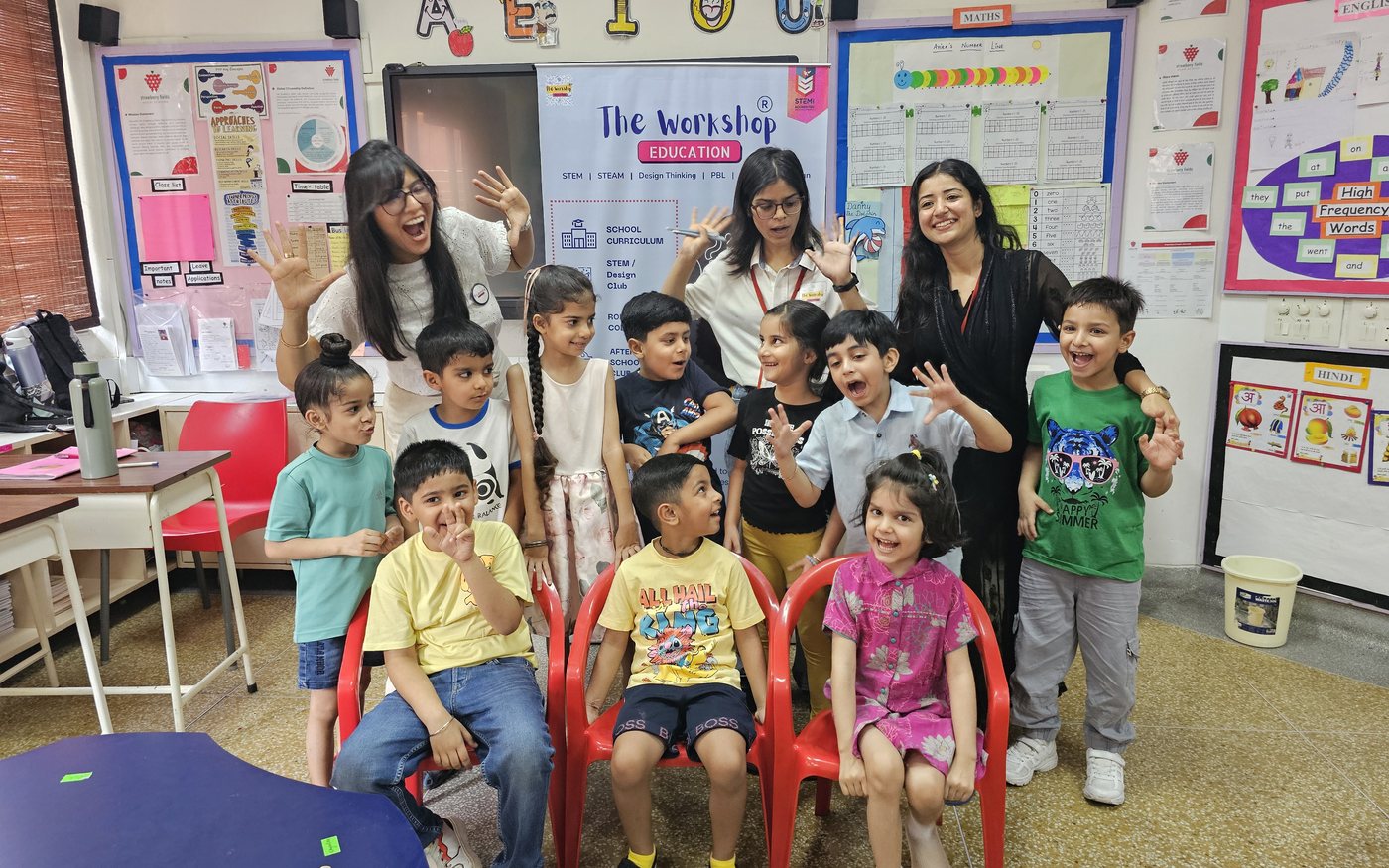 TheWorkshop educators and a group of young children posing together and laughing in a classroom, in front of a TheWorkshop Education banner, during a Holiday Club session.