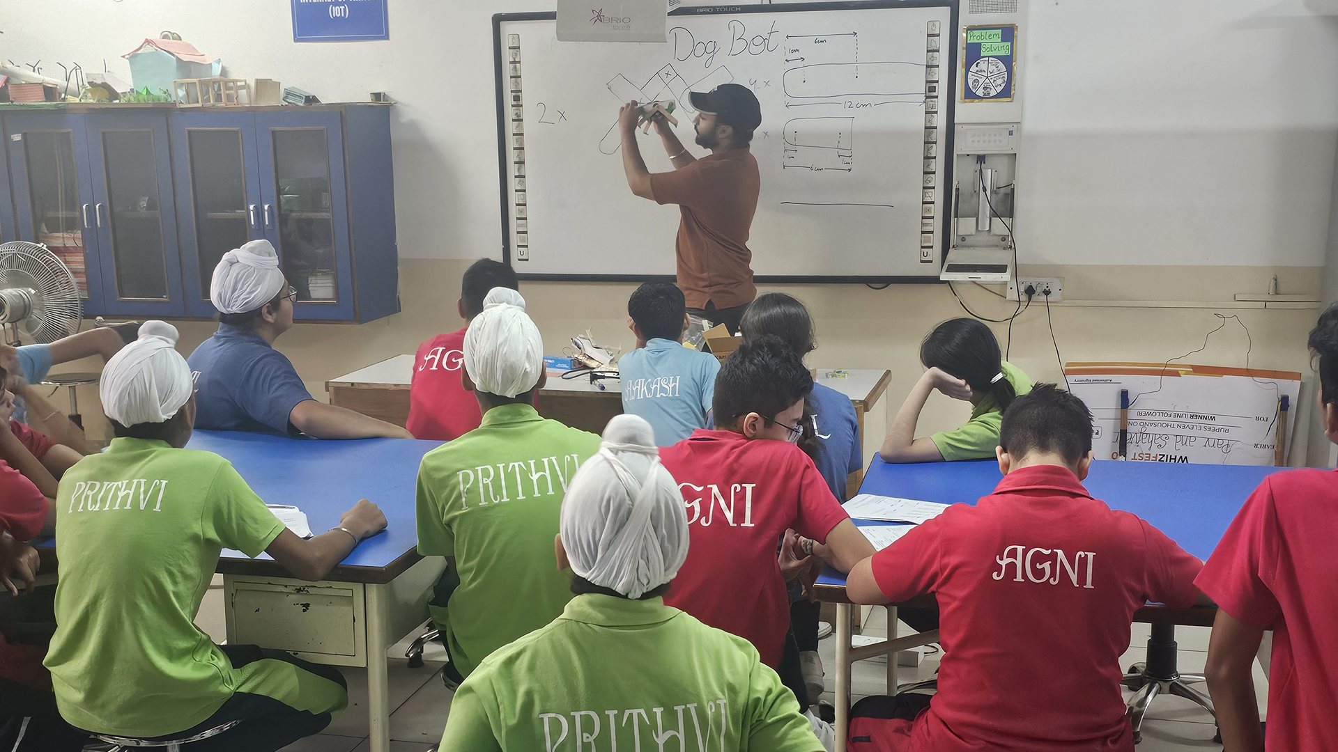 A TheWorkshop educator sketching a mechanism design on the whiteboard as middle school students watch, during an in-school STEM club session.