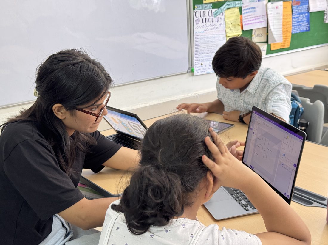 A tutor helping two students with laptop-based coding exercises at a table.