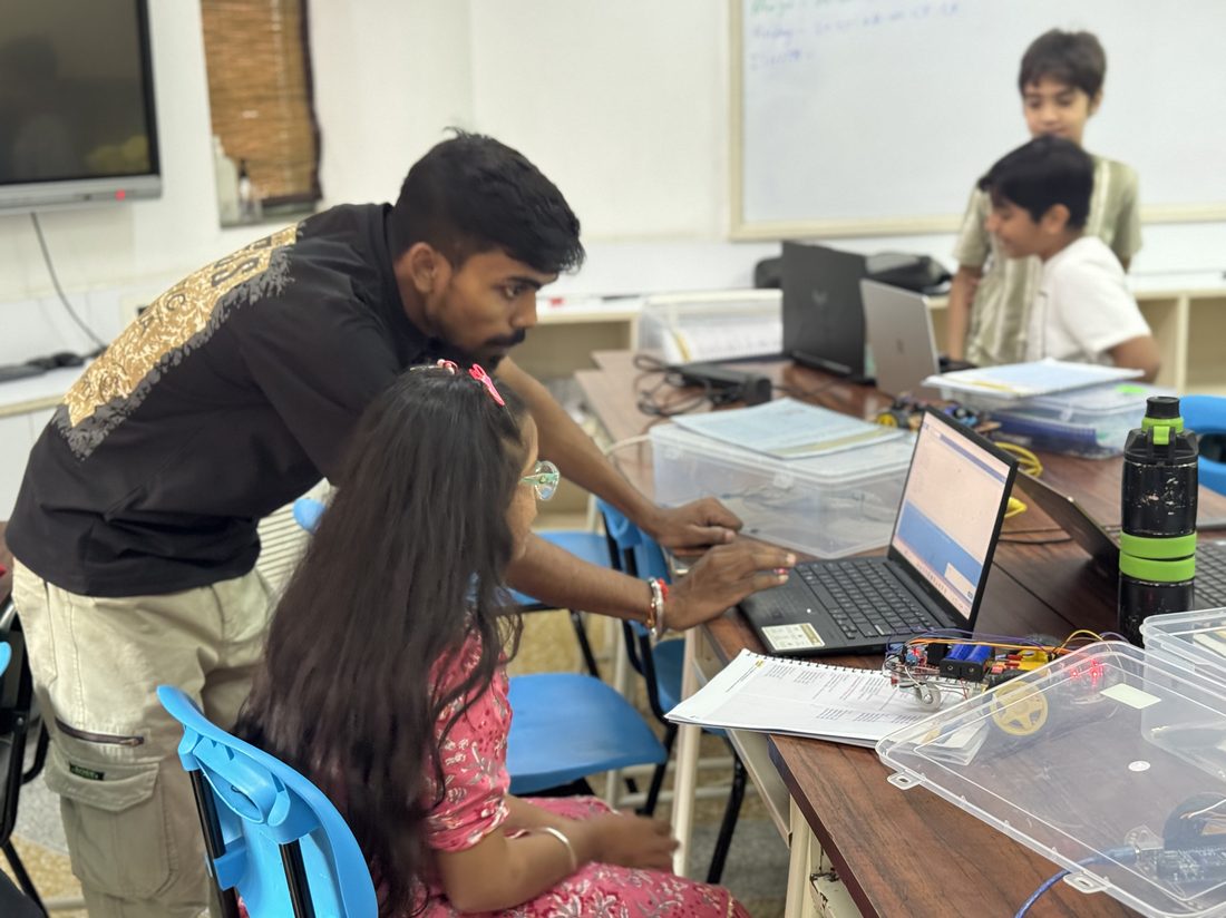 A mentor helping a student at her laptop, with other children working nearby.