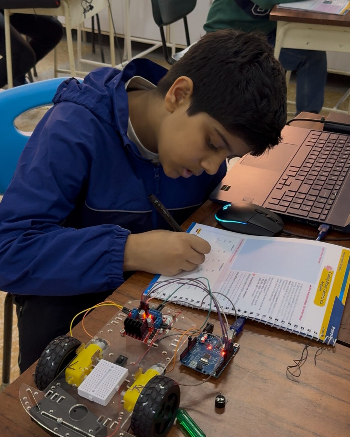 A student carefully wires an Arduino-based robot car, cross-checking the circuit against his lesson workbook.